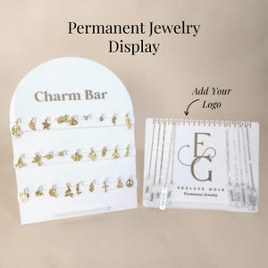 以下が含まれることがあります： 「Charm Bar」と「Permanent Jewelry Display」の文字が入った白いディスプレイスタンドに、金のチャームが飾られています。別のディスプレイには、ネームタグ付きのネックレスが展示されています。「Add Your Logo」の文字も表示されています。