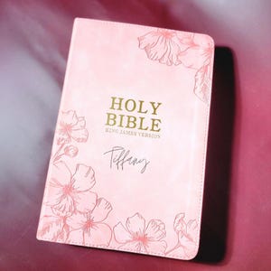 May include: A pink Holy Bible, King James Version, with floral illustrations and the name "Tiffany" in script. The cover features gold lettering and delicate flower drawings in the corners.