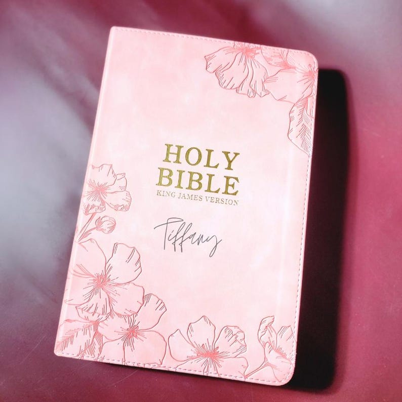 May include: A pink Holy Bible, King James Version, with floral illustrations and the name "Tiffany" in script. The cover features gold lettering and delicate flower drawings in the corners.