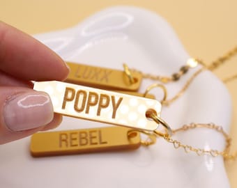 Puede incluir: Tres etiquetas de metal doradas con los nombres "Poppy", "Rebel" y "Luxx" grabados en ellas. Las etiquetas están unidas a una cadena dorada.