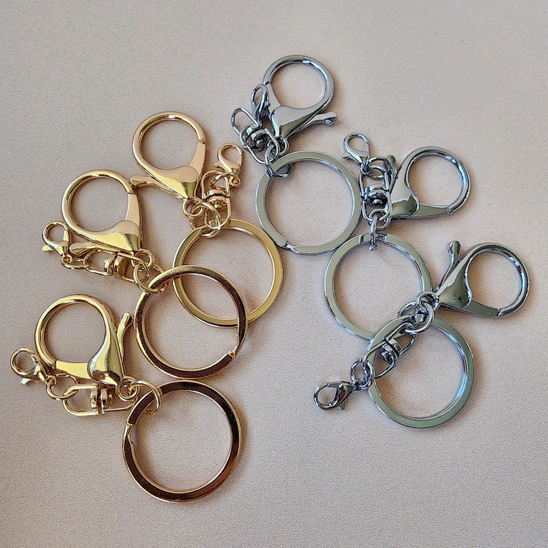 May include: An assortment of keychains in gold and silver tones. The keychains feature a variety of clasps, including lobster claw and swivel styles, attached to round key rings. The metal keychains are arranged on a neutral-colored surface.