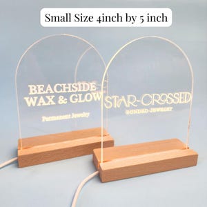 May include: Two acrylic signs with wooden bases, each with a different business name. The sign on the left says "BEACHSIDE WAX & GLOW Permanent Jewelry" and the sign on the right says "STAR-CROSSED BONDED JEWELRY". Both signs are 4 inches by 5 inches.