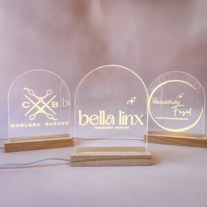 May include: Three illuminated acrylic signs with custom logos. The first sign says "Chelsea Barnes" with a logo of scissors and the letters "C" and "B". The second sign says "bella linx PERMANENT JEWELRY" with a star design. The third sign says "Beautifully Fused Custom Permanent Jewelry Bar" with a circle design.