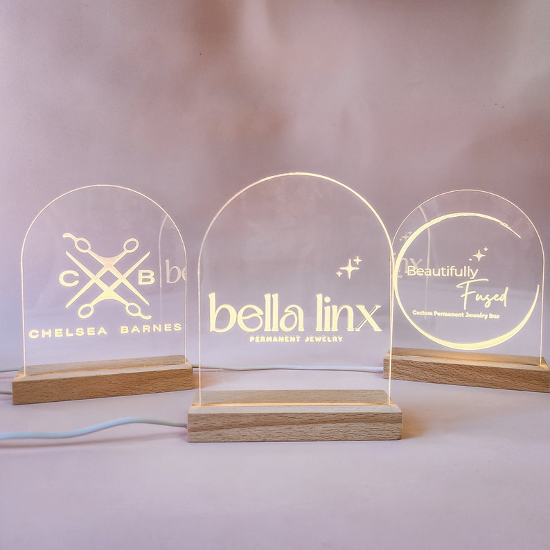 May include: Three illuminated acrylic signs with custom logos. The first sign says "Chelsea Barnes" with a logo of scissors and the letters "C" and "B". The second sign says "bella linx PERMANENT JEWELRY" with a star design. The third sign says "Beautifully Fused Custom Permanent Jewelry Bar" with a circle design.