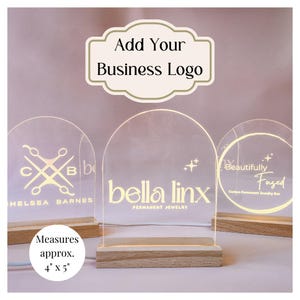 May include: Three illuminated acrylic signs with rounded tops and wooden bases. The signs feature different business logos and text, including "Chelsea Barnes", "bella linx PERMANENT JEWELRY", and "Beautifully Fused Custom Permanent Jewelry Bar". The signs measure approximately 10 cm by 13 cm.