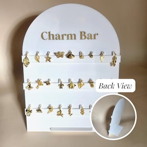 Puede incluir: Un expositor blanco con el texto dorado "Charm Bar" exhibe varios abalorios dorados. Los abalorios incluyen hojas, una estrella de mar, una concha y otras formas decorativas. El expositor tiene tres filas horizontales para exhibir los abalorios.