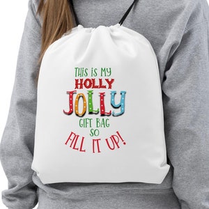 May include: White drawstring gift bag with the text "This is my Holly Jolly Gift Bag so Fill it Up!" in colorful letters. The bag has black drawstrings and is being worn as a backpack.