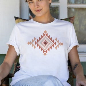 May include: A white t-shirt with a geometric design in shades of brown and pink. The design features a diamond shape with triangles and squares.