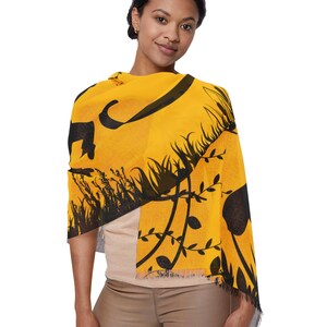 May include: A yellow scarf with a black floral design. The scarf is draped over the shoulders and has a fringed edge. The design includes silhouettes of flowers, grass, and other natural elements.