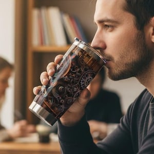 Puede incluir: Un vaso de acero inoxidable con un detallado diseño steampunk marrón y bronce. El vaso lo sostiene una persona en un café, con un portátil y un cuaderno sobre la mesa.