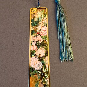May include: Gold metal bookmark with a floral design featuring pink and white flowers, green leaves, and a gold border. The bookmark has a blue tassel and the text "Van Gogh" at the bottom.