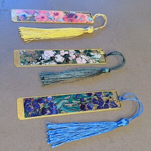 May include: Set of three gold-toned bookmarks with colorful floral designs and tassels. Each bookmark features a different floral painting and the artist's name, "Van Gogh", is printed on the bottom. The tassels are yellow, green, and blue.