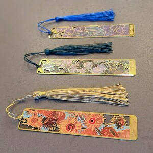 May include: Four gold-colored metal bookmarks with colorful floral designs and tassels. The bookmarks feature different floral patterns and include a tassel in blue, green, beige, and gold.