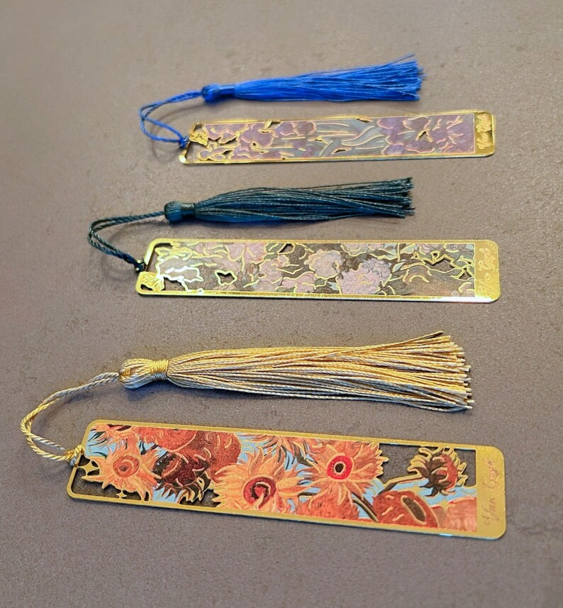 May include: Four gold-colored metal bookmarks with colorful floral designs and tassels. The bookmarks feature different floral patterns and include a tassel in blue, green, beige, and gold.