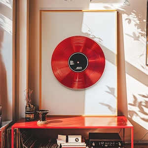 Könnte beinhalten: Eine rote Schallplatte mit einem schwarzen Etikett, auf dem "ADIO" steht, ist in einem goldenen Rahmen auf einem roten Regal ausgestellt. Die Schallplatte befindet sich an einer weißen Wand, durch die Sonnenlicht durch ein Fenster scheint.