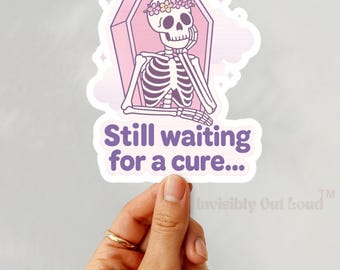 May include: A pink and purple sticker with a cartoon skeleton wearing a flower crown sitting in a coffin. The text on the sticker reads "Still waiting for a cure..." and "Invisibly Out Loud™".