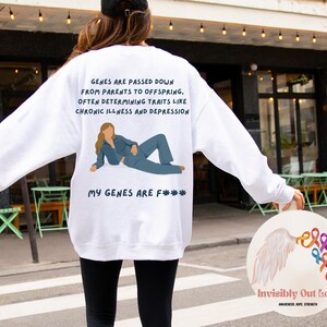 Könnte beinhalten: Weißes Sweatshirt mit Text: "GENE WERDEN VON ELTERN AN NACHKOMMEN WEITERGEGEBEN, OFT WERDEN EIGENSCHAFTEN WIE CHRONISCHE KRANKHEITEN UND DEPRESSIONEN BESTIMMT." Darunter eine Illustration einer Person in blauer Kleidung. Der Text "MY GENES ARE F***" steht darunter.