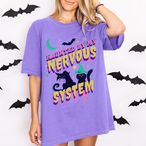 Può includere: Una maglietta viola con la scritta "HAUNTED BY MY NERVOUS SYSTEM" in giallo e viola. La maglietta presenta una luna crescente verde, un pipistrello nero e due gatti neri, uno con un cappello da strega verde. Pipistrelli neri decorano lo sfondo bianco.