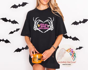 May include: Black t-shirt with a skeleton hand heart design and the text "Sick of Being Sick & Tired". The person is holding an orange Halloween mug. Black bat decorations are on the white wall behind the person.