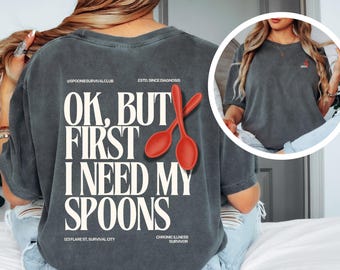 May include: Dark gray t-shirt with the text "OK, BUT FIRST I NEED MY SPOONS" and two red spoon graphics. The shirt also includes the text "SPOONIESURVIVALCLUB" and "CHRONIC ILLNESS SURVIVOR".