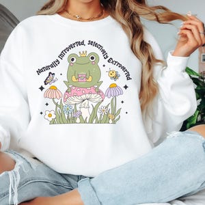 May include: White sweatshirt with a whimsical graphic of a frog wearing a crown, sitting on a mushroom, with the text "Naturally Introverted, Selectively Extroverted." The design includes a butterfly, bee, and flowers.