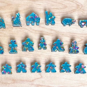 May include: A collection of teal-colored game pieces with iridescent glitter accents, arranged on a light wood board. The pieces are various shapes, including a house, cat, and people. The board is rectangular.