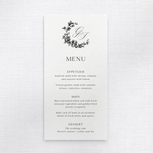Op de afbeelding: Een formeel menukaartje met een bloemenkrans en initialen bovenaan. Het menu bevat voorgerechten, hoofdgerechten en dessertopties, waaronder zeevruchtensalade, miso-kabeljauw en bruidstaart. De kaart is gedrukt op wit papier.