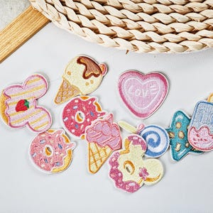 May include: A collection of embroidered patches featuring various dessert designs. Includes patches of ice cream cones, donuts, a heart-shaped candy with the word "LOVE", and a popsicle. The patches are in shades of pink, yellow, and blue.
