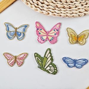 May include: Six embroidered butterfly patches in various colors, including blue, pink, green, and gold. Each patch features detailed stitching and a gold outline. The butterflies are arranged on a white surface, with a wooden frame and woven mat in the background.