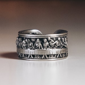 Può includere: Un anello d'argento con una rappresentazione dettagliata dell'Ultima Cena. L'anello è in metallo e ha una finitura liscia e lucida.