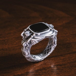 May include: A silver ring with a black gemstone and intricate details. The ring is set on a wooden surface.