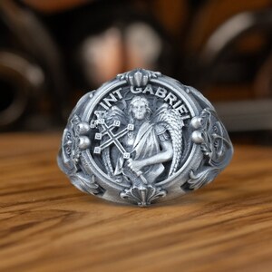 May include: A silver ring with a detailed design featuring an angel holding a cross. The ring is inscribed with "SAINT GABRIEL".