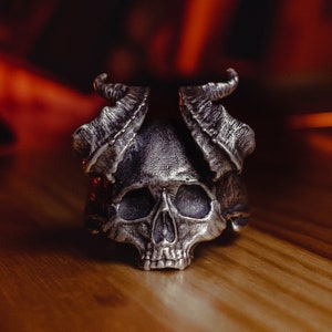 May include: A silver skull ring with horns, the skull has a detailed design and is set against a blurred orange background.