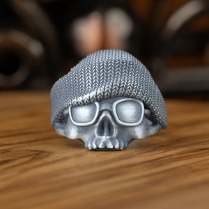 Op de afbeelding: Een zilveren schedelring met een grijze gebreide beanie erop en een zonnebril. De ring ligt op een houten oppervlak.