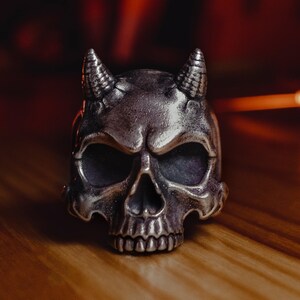 May include: A silver skull ring with horns, the skull has a detailed design and is set against a wooden background.
