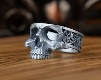 May include: A silver skull ring with intricate floral details on the band. The ring is lying on a wooden surface.