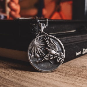 May include: A silver pendant necklace with a detailed design of a bird in flight. The pendant is round and has a textured surface. The necklace is resting on a black notebook.