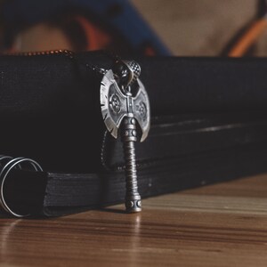 May include: A silver axe pendant with intricate details hangs from a black chain. The pendant is resting on a black notebook with a spiral binding.