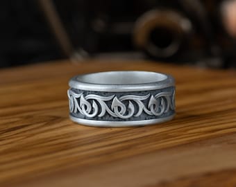 May include: A silver ring with an engraved floral pattern. The ring is set on a wooden surface, with a blurred background. The ring's design features a repeating pattern of stylized leaves and swirls, creating a textured band.