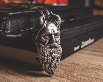 Puede incluir: Un collar con colgante plateado que representa a un hombre barbudo con cuernos. El colgante cuelga de una fina cadena y descansa sobre un libro encuadernado en cuero oscuro. El fondo incluye una superficie de madera y una mano borrosa.