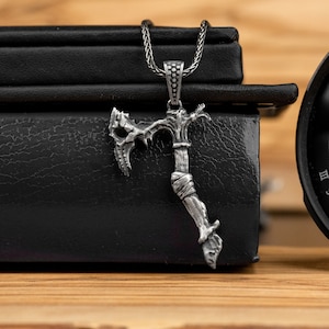 以下が含まれることがあります： 詳細なデザインの銀製の斧のペンダントネックレス。ペンダントは銀のチェーンにぶら下がっており、黒い表面の上に置かれています。