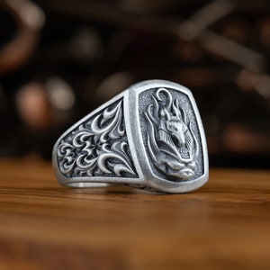 Puede incluir: Un anillo de plata con un diseño intrincado que presenta una cabeza de lobo con llamas. El anillo tiene una banda ancha con un patrón floral detallado.