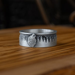 May include: A silver ring with a wolf howling at the moon design. The ring features a detailed wolf head and a forest scene with pine trees.