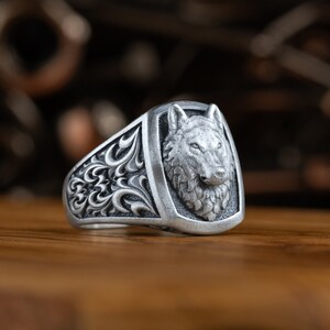 May include: A silver ring with a detailed wolf head design. The ring has a textured band with intricate floral patterns.