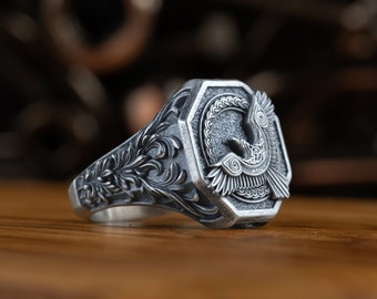 May include: A silver ring with an intricate design featuring a bird with outstretched wings. The ring has a detailed floral pattern on the band.