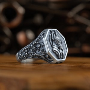 Puede incluir: Un anillo de plata con un diseño intrincado que presenta una representación detallada de una cabeza de animal. El anillo tiene un bisel de forma octogonal y una banda con patrones florales.