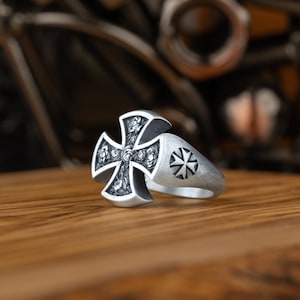 Könnte beinhalten: Ein silberner Ring mit einem Kreuzdesign mit fünf Rosen. Das Kreuz ist mit einer strukturierten Oberfläche verziert und der Ring hat ein Sternendesign.