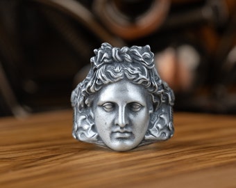 May include: A silver ring with a detailed sculpted face of a Greek god or goddess. The ring is on a wooden surface.