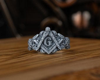 May include: A silver ring featuring an intricate design of a compass and square, with a letter 'G' at its centre. The ring is displayed on a wooden surface.