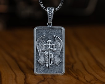 May include: Silver pendant necklace featuring a rectangular charm with a detailed 3D design of an angel holding a sword. The angel has wings and is kneeling.